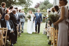 The bride walks down the "aisle" between the 2 colums of wooden chairs that are set out in front of the arbour. SHeis on her fathers arm. She is on the left walking towards the camera. She wears a full length white, sleeveless dress with lace details. She wears a veil. She has a bouquet of flowers, mostly green, in her right hand. The guests are stannding and turning around to see her.