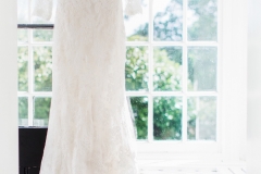 Lace, white wedding gown hanging in the porch of the the house. In the background there are Georgian say windows and a black painted front door that is open.