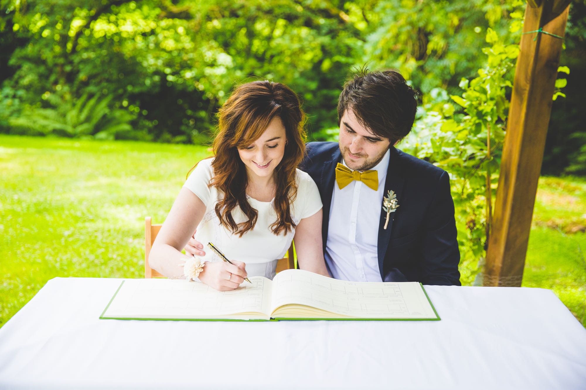 This image shows an elopement couple signing the register. The couple are seted at a white clothed table. In front of them is the open register. The bride sits on the left and the groom on the right. They are both looking at the book and the bride has a pen in her right hand. The groom has his arm around the bride's waist. The bride is wearing a cap-sleeved, white dress and has a flower on her wrist. She has long, dark hair that she's wearing loose. The groom is wearing a dark blue jacket, a white shirt and a yellow bowtie. He has dark hair and a beard. In the background there are lots of green trees, shrubs and grass. On the right you can see one of the oak posts that form part of the Lake Arbour.