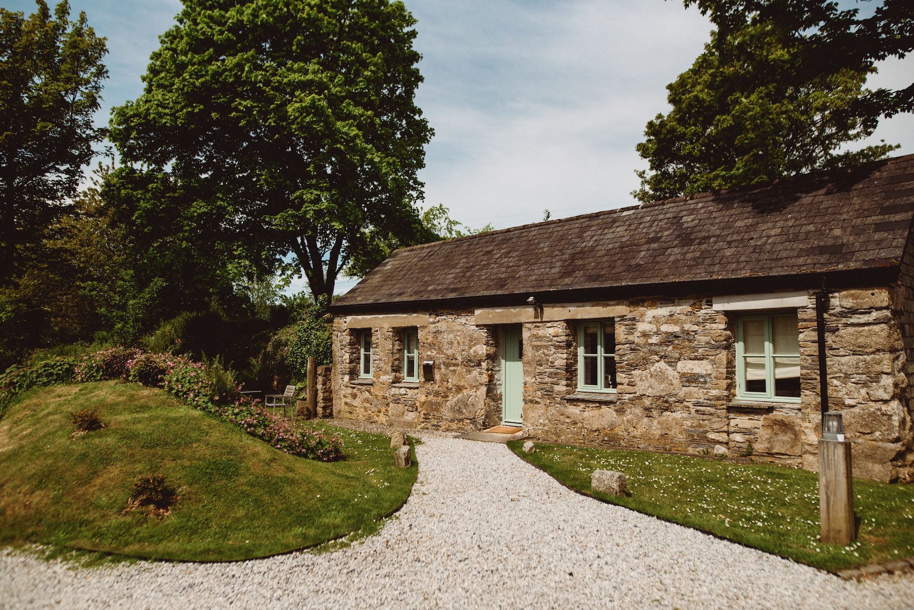 Cottage One which is a single storey building with two windows on either side of the door. All the paintwork is pale green and the stonework is original. There is a gravel path leading towards the door and a grassy bank on the left hand side and grass to the right. It is a sunny day and there is blue sky and light cloud in the background