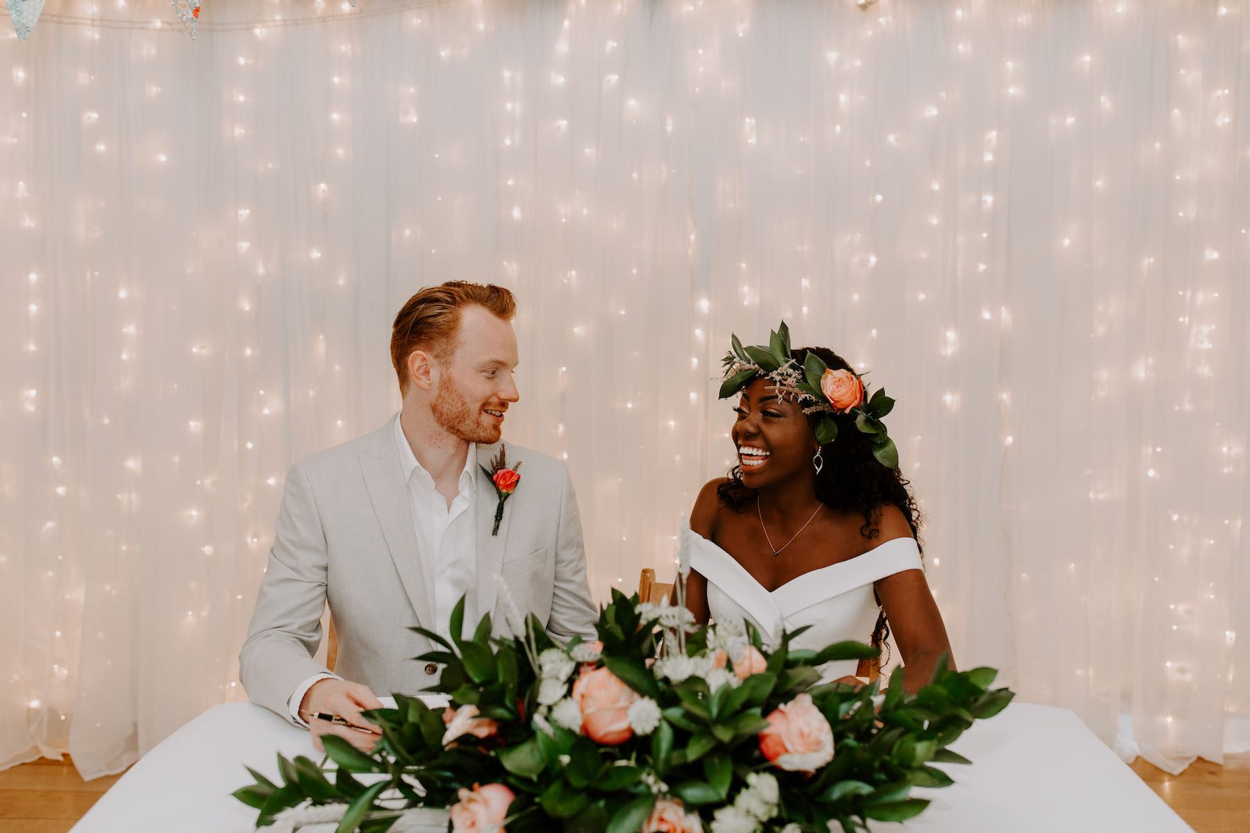 An elopement wedding with the bridal couple seated at the registration table in the Wedding Barn. There is an arrangement of pink roses on the table and a curtain of fairly lights behind the couple. The bride is wearing a headdress of natural roses and greenery and the groom is wearing a buttonhole of a pink rose on his left lapel