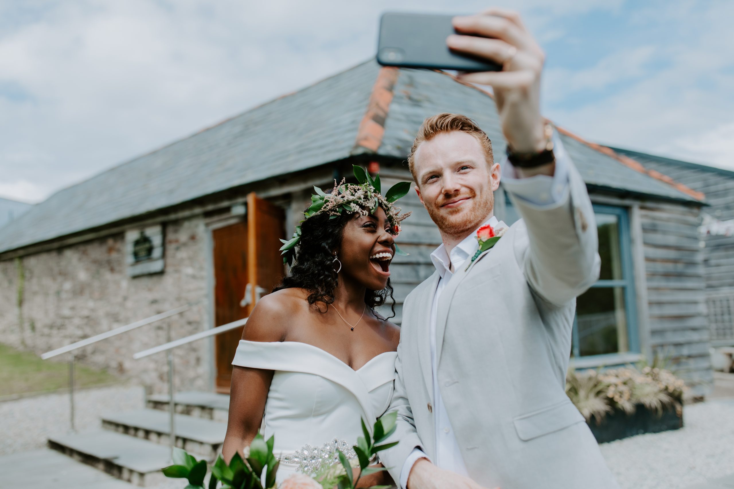 A bridal couple taking a selfie outside with the Wedding Barn in the background. The bride is wearing a white off-the-shoulder dress with diamente detail at the waist and a headdress of natural pink flowers and greenery. The groom is wearing a pale grey suit and a single pink rose buttonhole with an open neck shirt. They are both smiling at the camera.