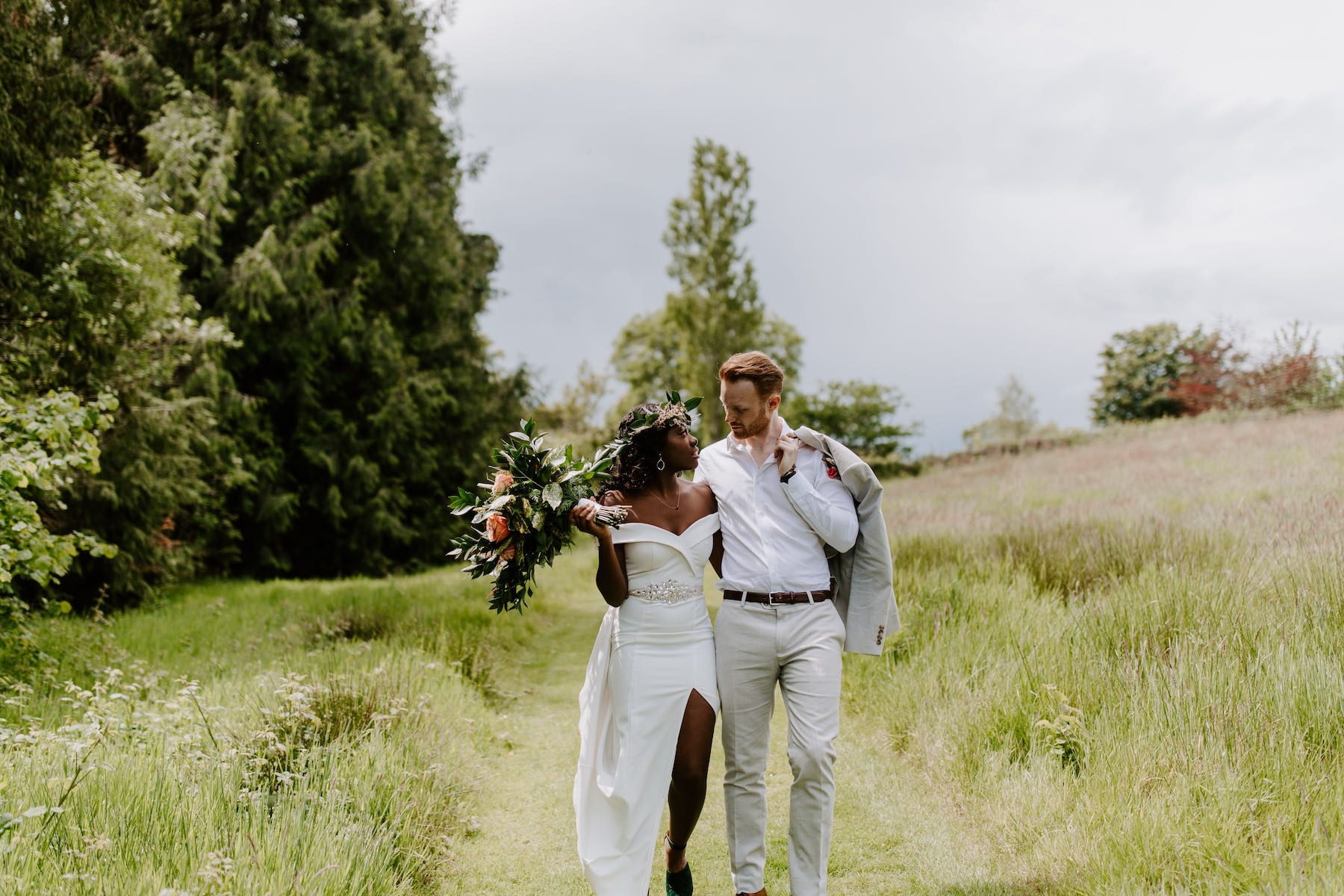 A bridal couple are strolling down the cut path of the meadow in summer with long grass on either side of the path and trees in the background. The bride is wearing a white dress with a slit up the left side and diamente detail at the waist. She is carrying a bouquet of pink roses and greenery which she is holding up towards her right shoulder and the groom who is wearing a light grey suit is holding the jacket on his left shoulder. They are looking at each other lovingly.