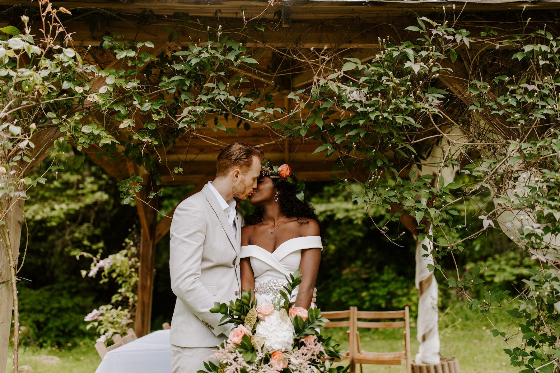 A bridal couple standing outside at the Lake Arbour, kissing. The Bride is wearing a white off the shoulder dress and holds a large bouquet of pink roses and greenery and the groom is wearing a pale grey suit. The couple are framed by the Arbour which is covered in trailing clematis leaves.