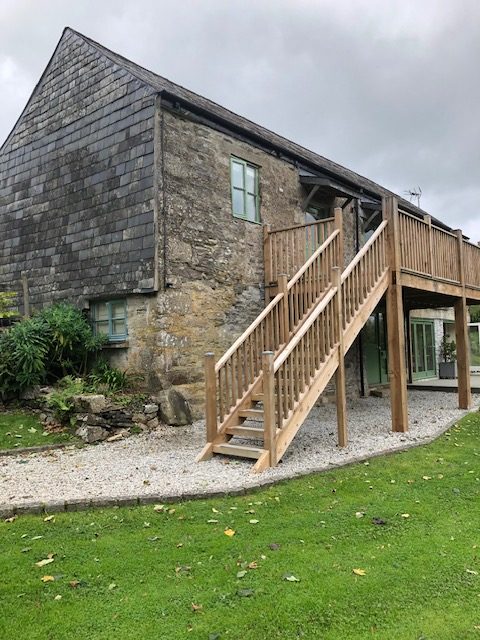 A two-storey stone cottage, a former agricultural building with wooden steps leading up to a balcony area giving access to two cottages on the first floor. Below another cottage with outside decking area. A gravel path leads to the steps and a grass lawn is situated in front of the building