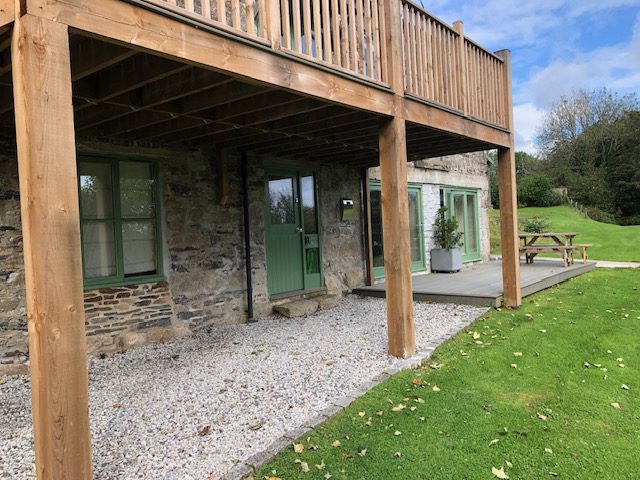 A ground floor stone building with green windows and doors framed by the wooden balcony structure for the upper level. A gravel path leads to the right of the building opening onto a decking area showing a picnic table and a planter with a flat lawn area stretching across the whole of the front of the building.