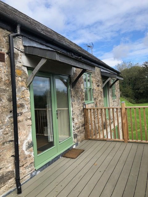 The entrance to one of the stone built cottages showing the decking space and surrounding wooden balustrades. The entrance door is pale green and glass with a window to the right and further unused door beyond.