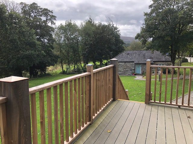 An open decking area with balustrades to the left and ahead with a gap where the steps lead down to ground level. In the distance ahead is a single stoney property with a grass lawn in front and a number of large leafy trees set against a cloudy sky