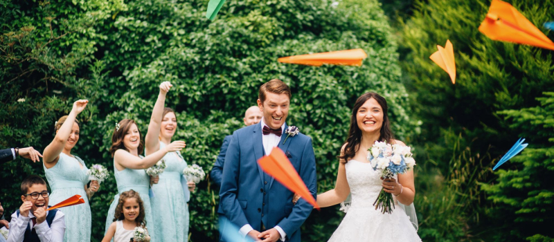 exclusive-hire-5 The image shows the bride and groom standing surrounded by trees with some guests around them. Their guests are throwing paper aeroplanes instead of confetti. The aeroplanes are in bright colours. There trees behind the couple who are standing. The trees are in full leaf. The image shows the couple from knees up. The bride is wearing a beaded, full-skirted, strapless dress. In her left hand she is holding a hand tied bouquet with blue and white flowers. She has dark, long hair that she's wearing down. The couple are facing the camera and smiling. The groom is wearing a light blue three piece suit with a brown bow tie and a white shirt. In the background you can see a little girl and little boy and three bridesmaids. The bridesmaids are wearing mint green, knee-length bridesmaids dresses. Their hands are raised as they throw the aeroplanes.