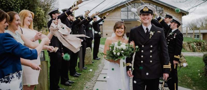 frances-ian-12 A couple walk away from the green room bar under the arch formed by swords. While they walk away their guests throw pom-pom confetti at them them. The bride is on the left and the groom is on the right. The groom where is a naval dress uniform. The bride wears a white, floor length gown with shoestring strap white and carries white and green flowers. The Green Room is a wooden chalet style building with glass frontage. This is an April wedding and the sky is overcast.