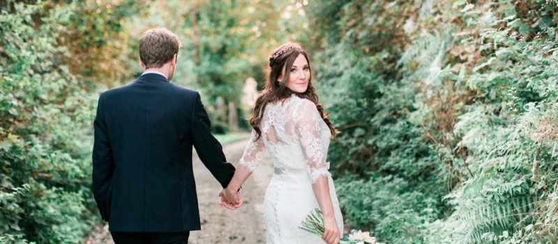 rural-romance-.0.5 The bride and the groom are walking away from he camera down a narrow and leafy Cornish lane. The bride is on the right. She wears a fitted white gown with lace details and a delicate floral crown. The pic is takes from the back and the bride is looking over her right shoulder back at the camera. The groom wears a dark suit. The bride holds a bouquet of white and pink flowers in her right and it is hand-tied.